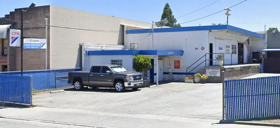 White building bulk plant with a blue roof and a gray truck parked out front.