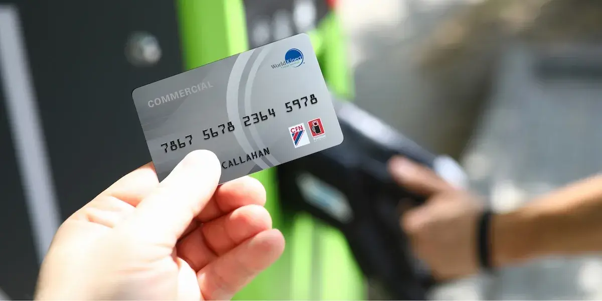 A close-up of a silver "Commercial" fuel card held by a hand. The card displays the World Kinect Energy Services logo in the top right. Small logos for the CFN and Fuelman networks are visible in the bottom right corner. In the background, a green and black fuel pump is out of focus.